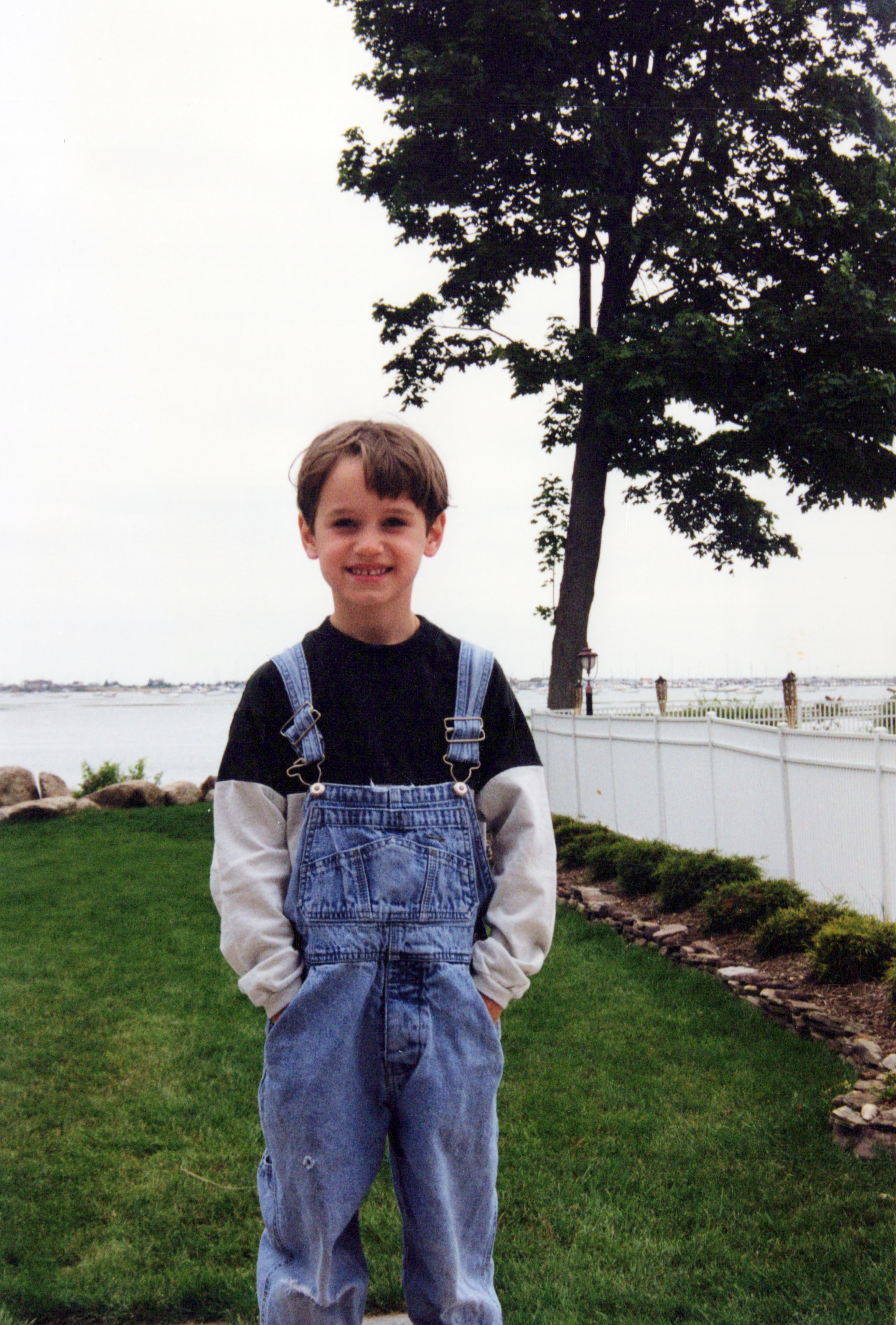 A young boy in denim overalls stands on a grassy lawn near a waterfront, late 1990s